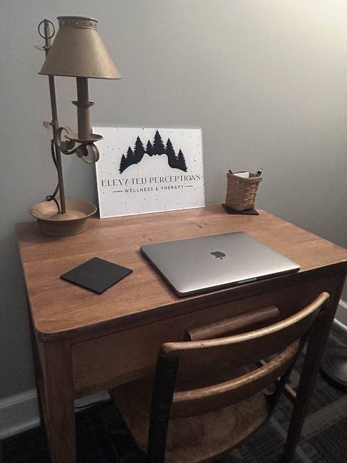 A small wooden desk with a laptop, a lamp and the Elevated Perceptions sign, set up for a telehealth session
